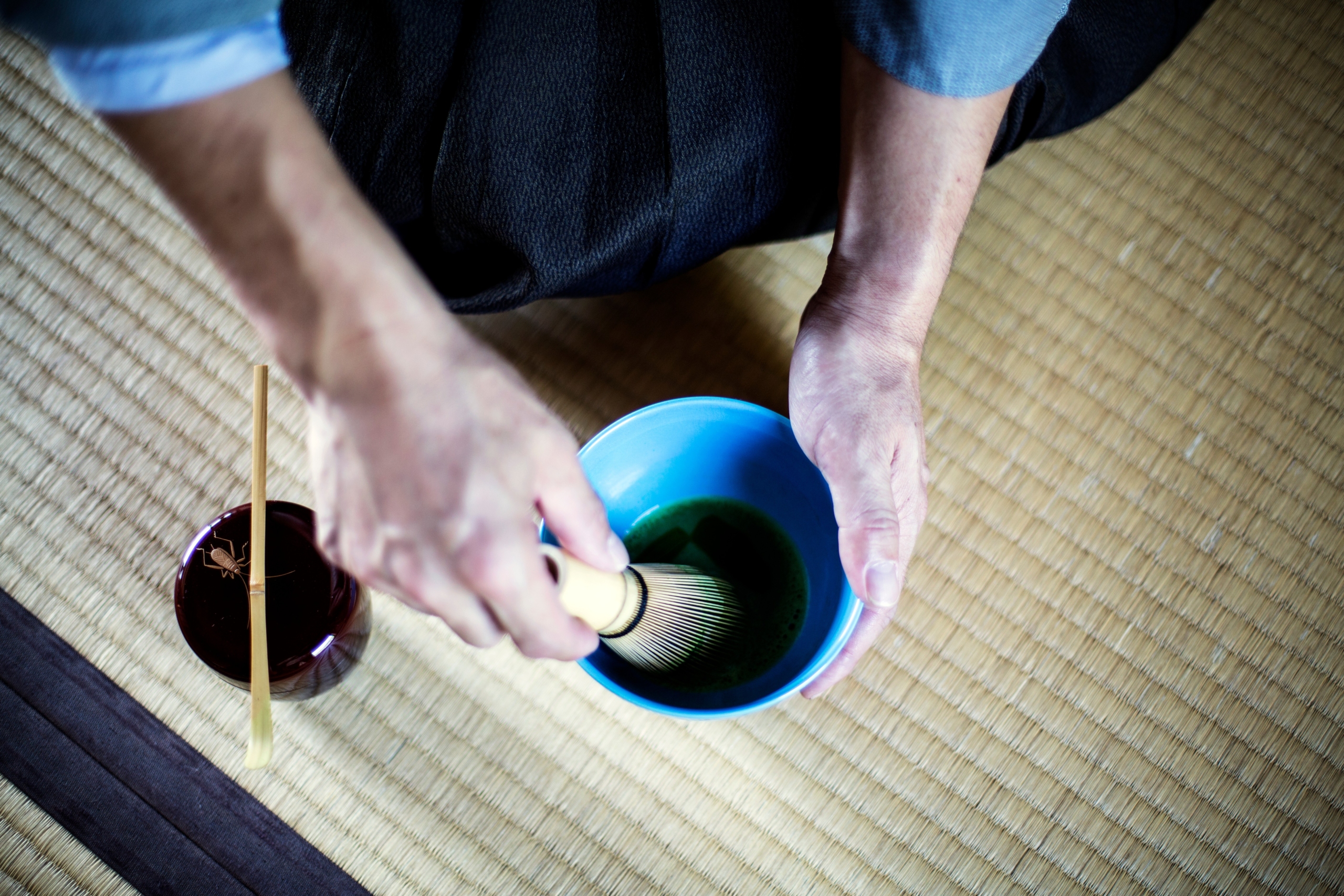 Japanese man using bamboo whisk to prepare Matcha tea in a blue bowl during tea ceremony.